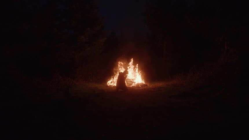 A solitary figure kneels in front of a large burning bonfire in a dark wooded clearing