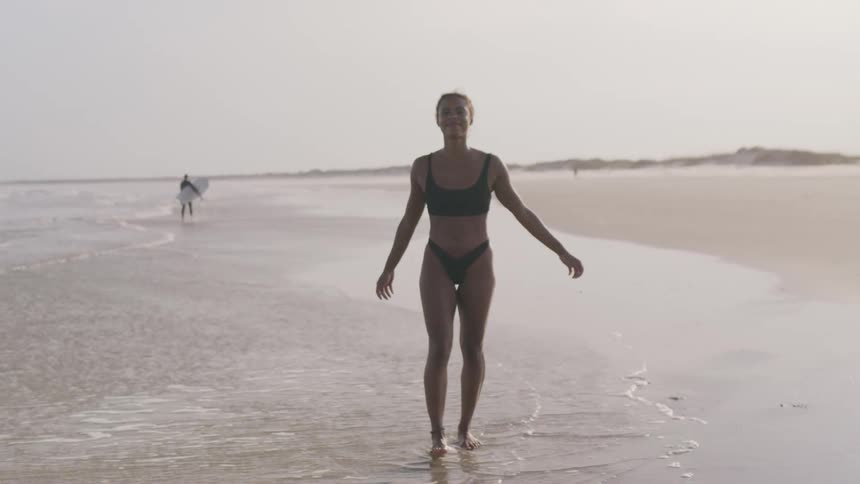 A woman in a dark bikini walks confidently toward the camera along the wet sandy shoreline as a surfer with a surfboard can be seen in the distance