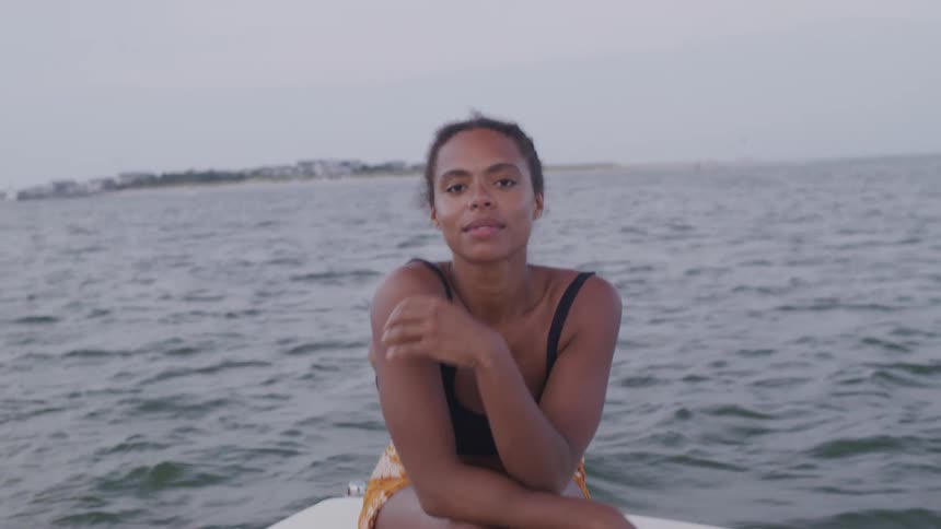 A woman with her hair blowing in the wind sits on the deck of a boat looking out at the water with a small smile on a sunny day