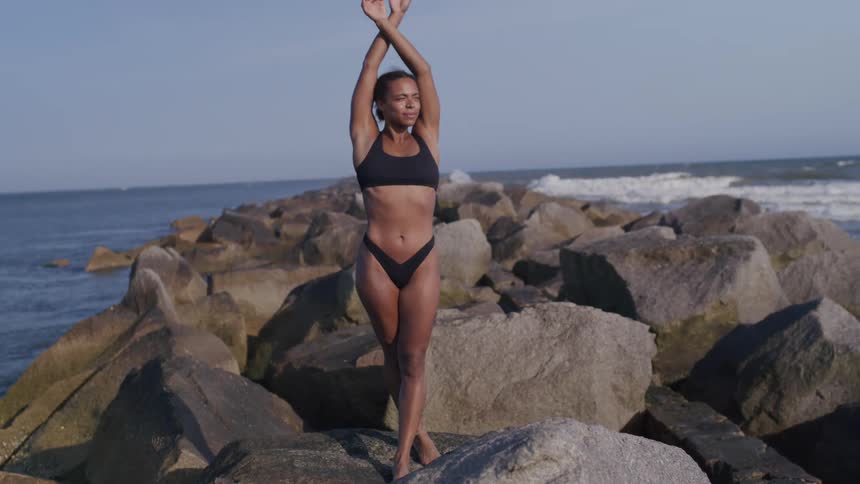 Woman in a bikini standing on rocks near the ocean with arms raised