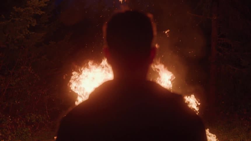 A man stands silently facing a towering blaze in the forest as sparks rise into the night sky