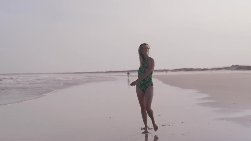A woman in a green and black one piece bathing suit walks and smiles towards the camera on a sandy beach