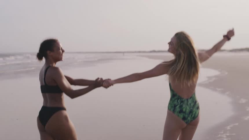 A woman in a black bikini and a woman in a green and black one piece bathing suit walk hand in hand down a wet sandy beach smiling and interacting with one another