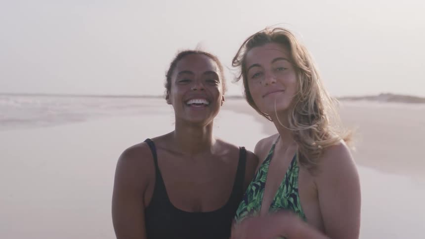 Two women hug each other on the beach wearing swimsuits at midday smiling