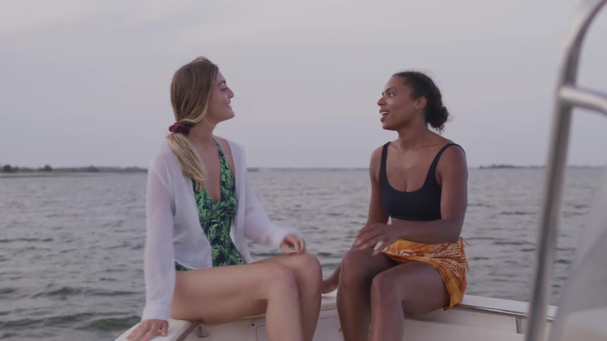A wide shot of two women sitting on a boat laughing and enjoying a conversation on the open water