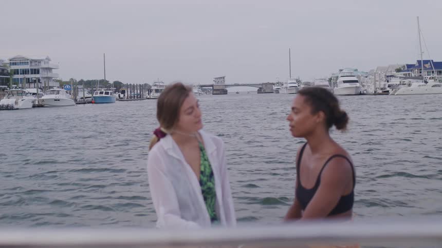 Two women sit and chat on a boat in a marina surrounded by calm water and docked yachts