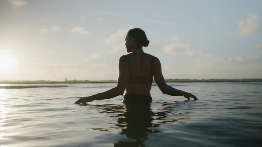 A woman with a bun in her hair and a dark top wades in the ocean with her back to the camera looking to the side as the sun sets behind her