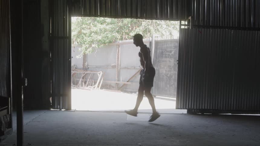 A man is exercising by jumping rope in a warehouse or gym the warehouse doors are open revealing trees and a bright day outside