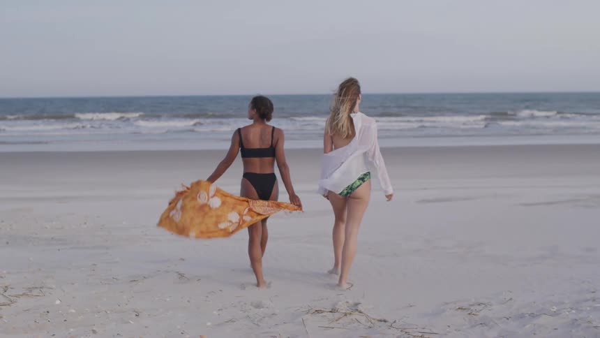Two women walk barefoot on the beach toward the ocean one holding a flowing orange scarf