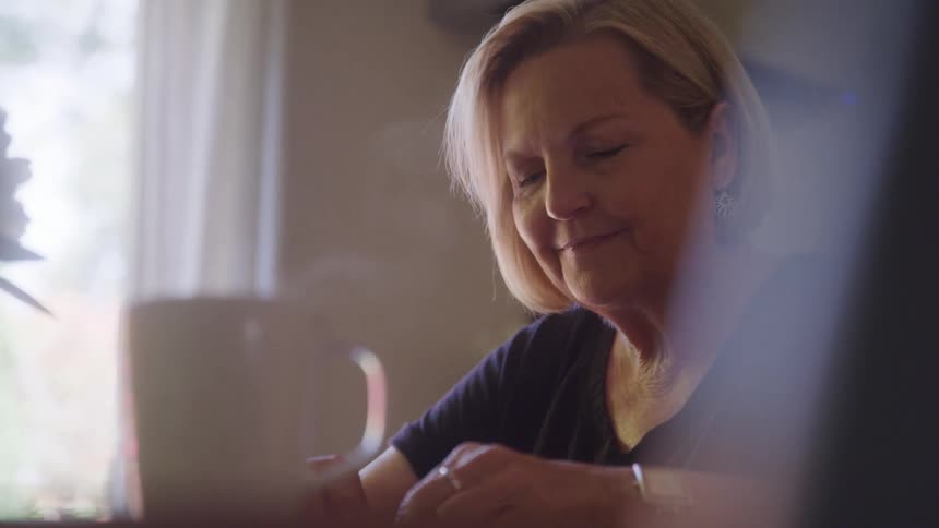 A middleaged woman with blonde hair is shown in a closeup focused on doing something as soft light from a nearby window illuminates the scene