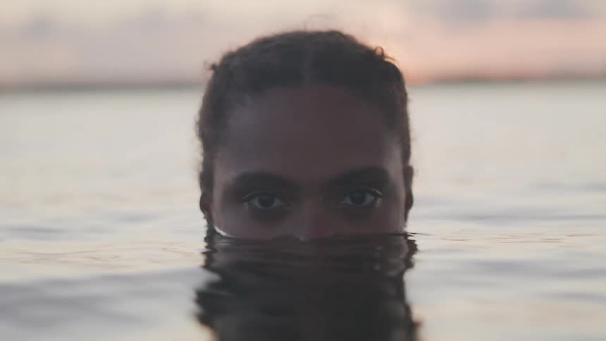 A woman emerges from water at sunset with only her eyes and forehead visible above the surface