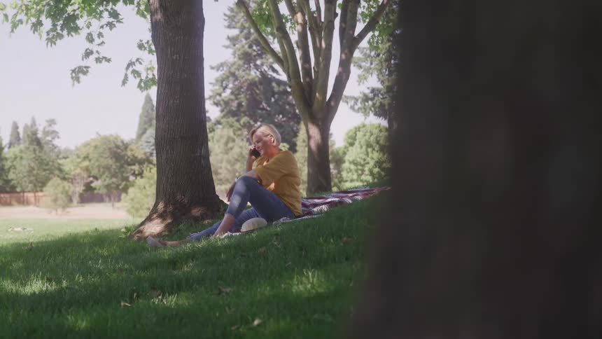 A woman in a yellow sweater and jeans sits on a picnic blanket on a grassy hill under the shade of a tree talking on her cell phone
