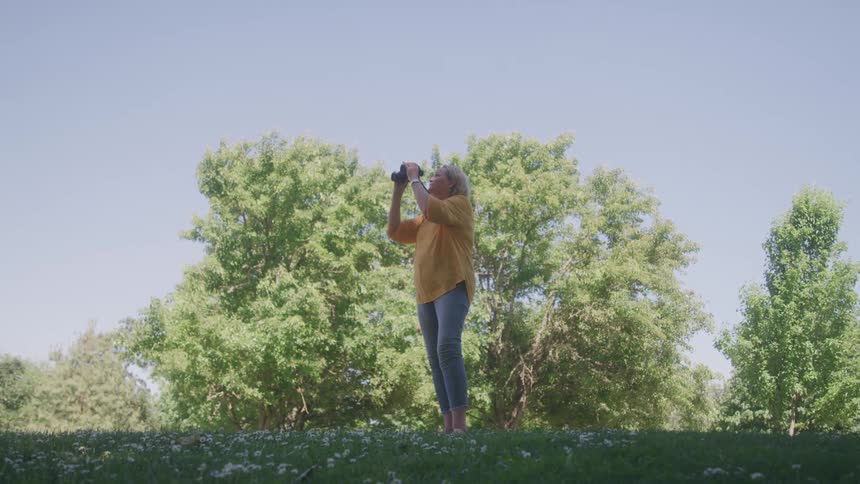 An elderly woman on a grassland uses binoculars for birdwatching with a smile on her face at midday