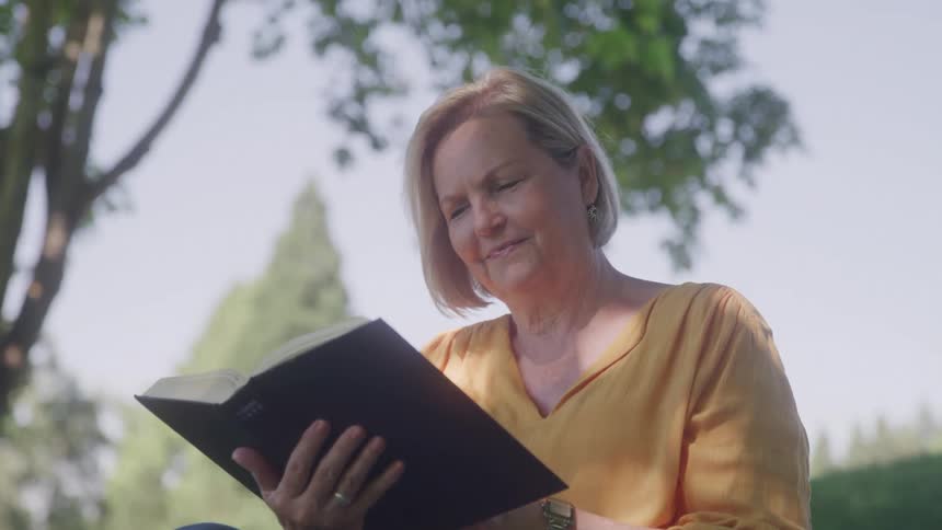 Woman in a yellow blouse smiles while reading a book outdoors