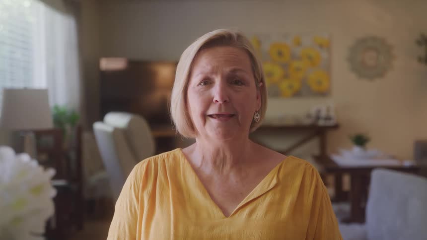 The video shows a woman with short blonde hair and a yellow shirt smiling and waving directly at the camera the setting appears to be a living room with a dining area and a large window in the background