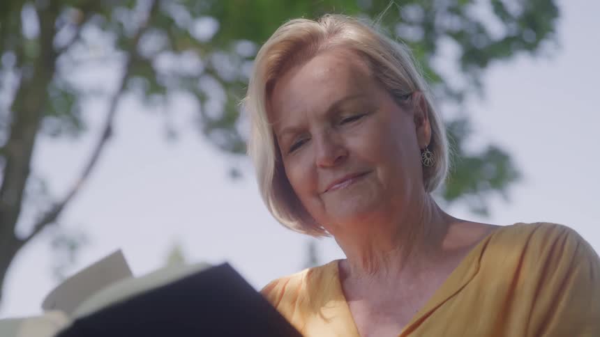 A woman with short blonde hair reads a book outdoors under the shade of leafy trees