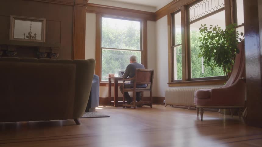 A man sits alone at a wooden table by a large window in a sunlit living room