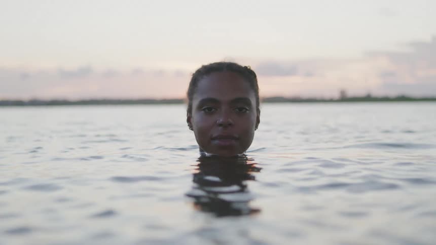 A woman emerges silently from the water at sunset with only her eyes and forehead visible above the surface