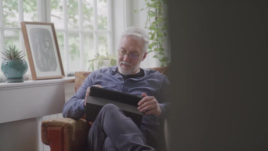 A person with gray hair and glasses is sitting in a brown armchair holding a tablet sunlight is coming through a window behind him and there are plants and a framed picture on the windowsill