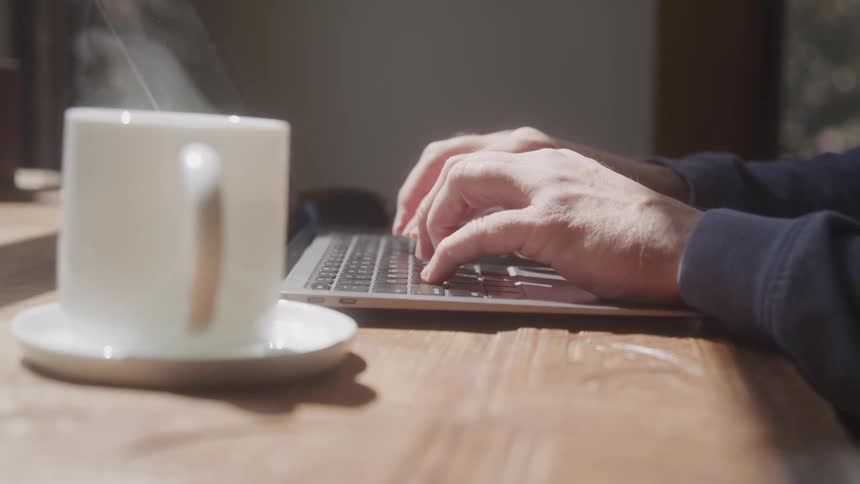 A person types on a laptop at a sunlit wooden table beside a steaming white coffee mug