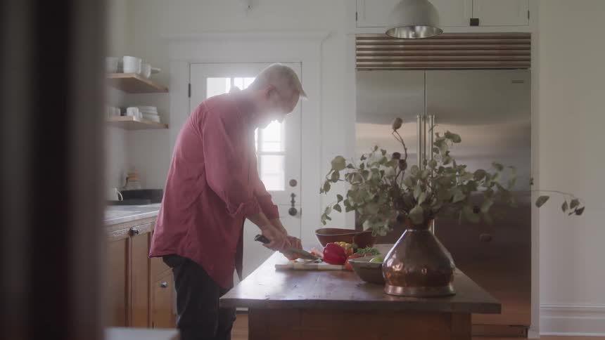 A person with gray hair wearing a red shirt is standing at a wooden kitchen island chopping vegetables in the foreground a large copper vase with dry leaves or branches sits on the island partially obscuring the view