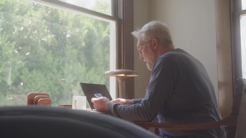 A man sits at a desk in a welllit room looking at a laptop screen while holding a credit card and appearing to make an online purchase