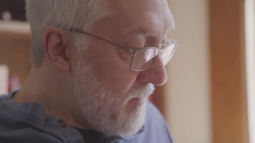 A closeup profile shot of an elderly man with a white beard and glasses looking down with a focused and thoughtful expression