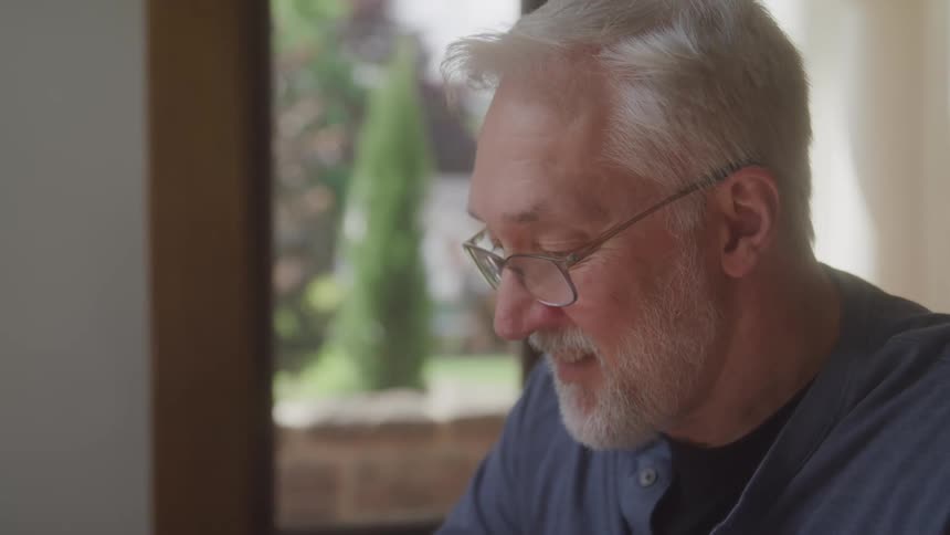 A bearded man in glasses smiles quietly while looking downward in a sunlit room with plants nearby