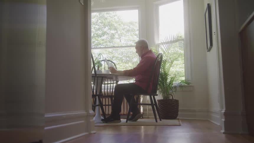 A man in a red shirt sits at a small table near a bright window with potted plants nearby