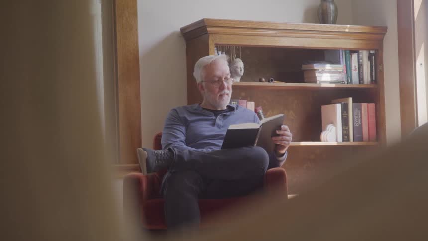 Older gentleman sitting on a chair near a window reading a book