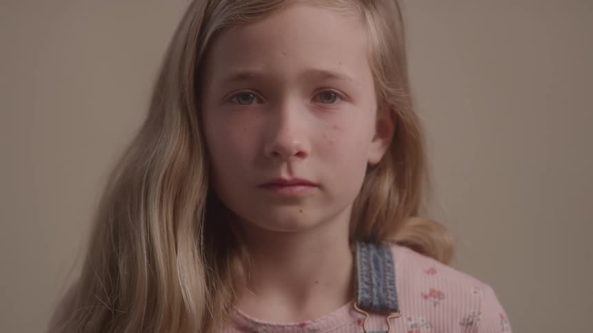 A young girl with long blonde hair gazes solemnly into the camera against a neutral backdrop