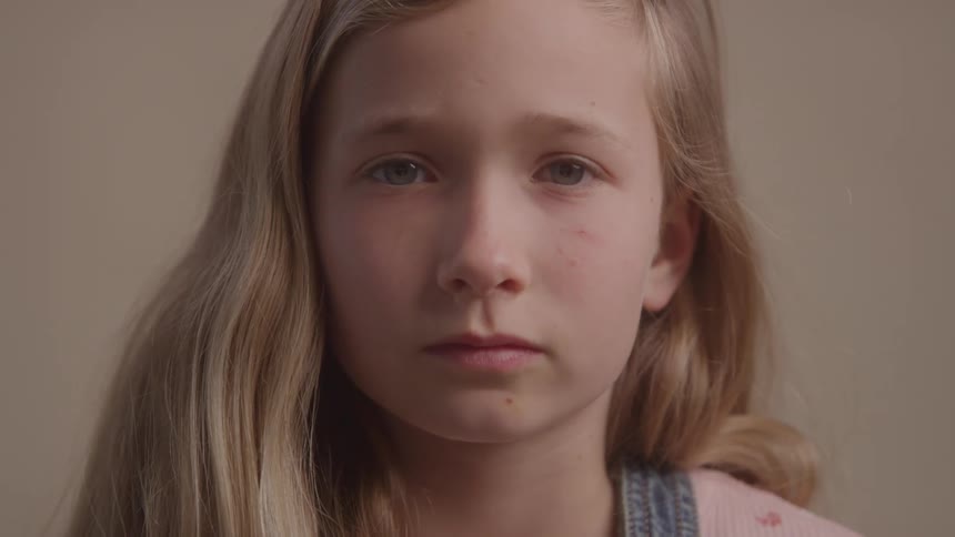 A young girl with long blonde hair and blue eyes looks directly at the camera with a serious expression