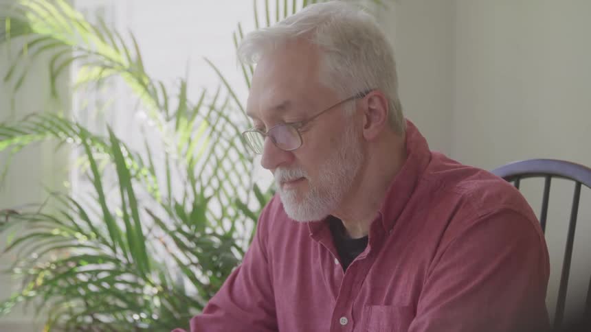 An older man in a red shirt and glasses sits near a window focused on looking downward