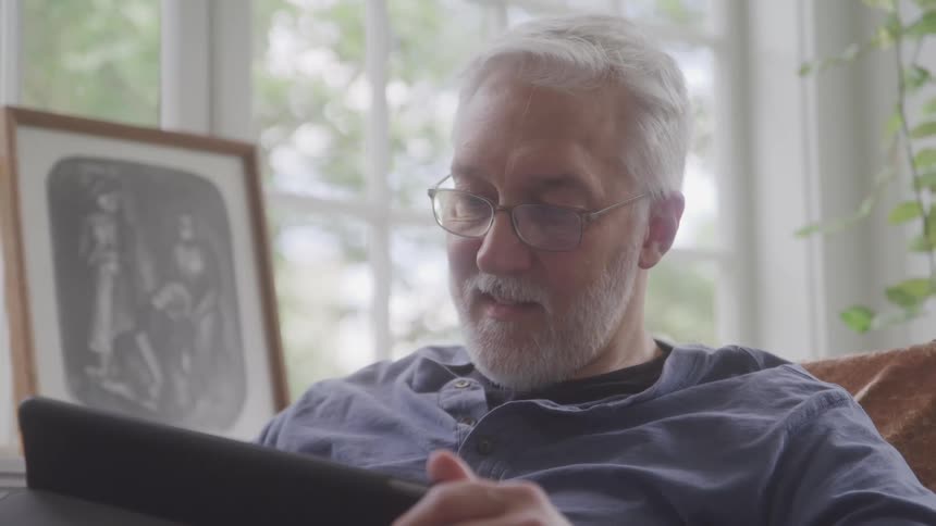 A man with a white beard and glasses sits on a couch holding a tablet and smiling as he looks at the screen