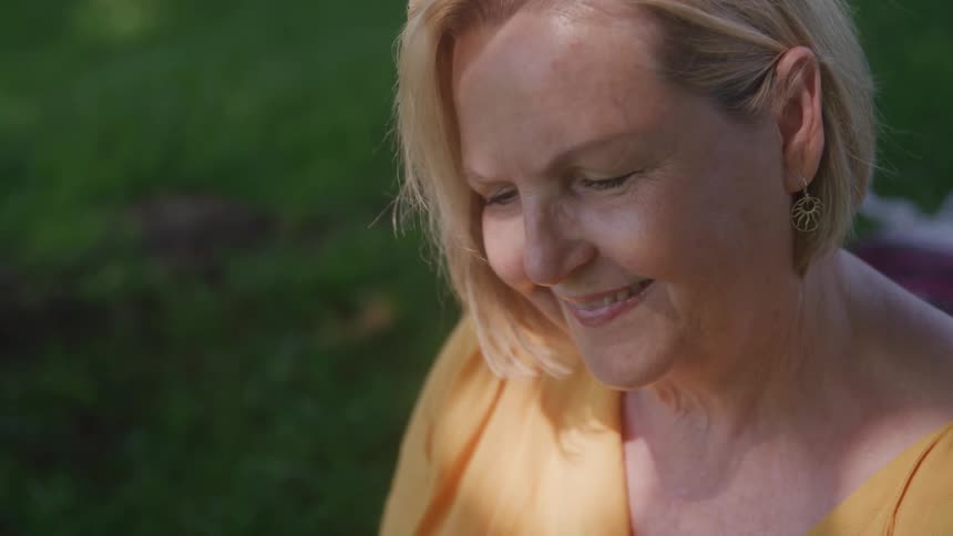 The video shows a closeup of a middleaged woman with short blonde hair she is smiling softly while looking down and to the side with a blurred green background suggesting she is outdoors in a park or garden
