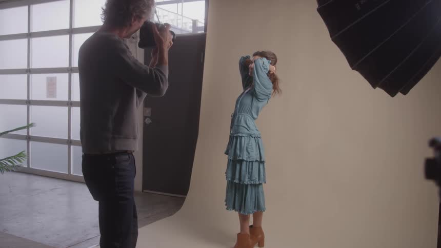 A photographer is taking pictures of a model in a studio the model is wearing a long blue tiered dress and posing with her hands near her head