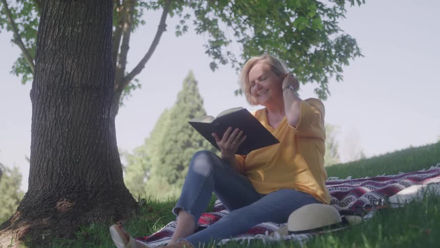 A woman is sitting on a red and white checkered blanket under a large tree reading a book with a happy expression on a sunny day