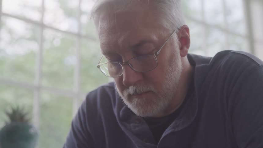 Man with glasses reads intently near a bright window