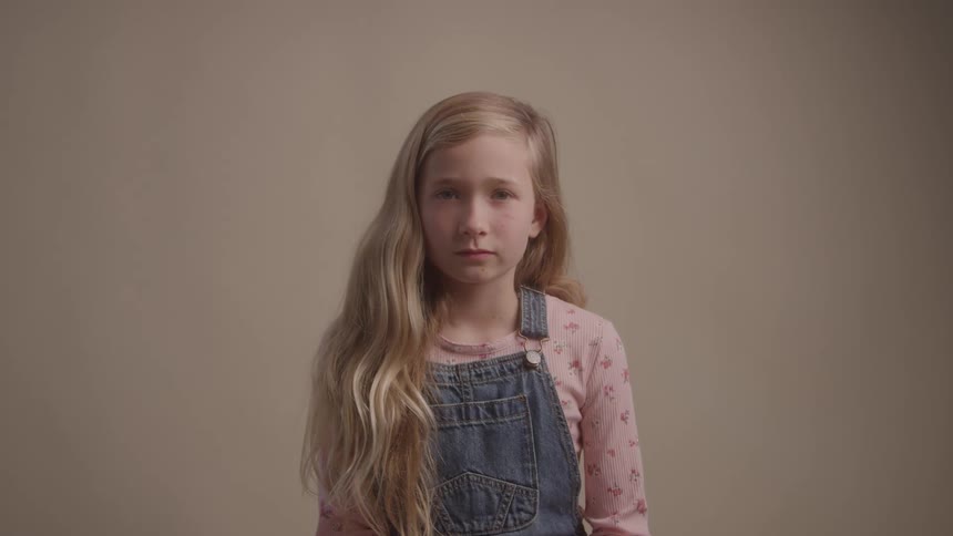 Blonde girl with long hair wears a pink shirt and denim overalls against a neutral background