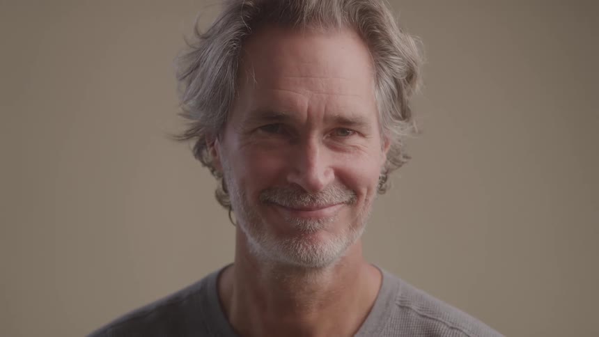 A man with gray hair and a relaxed smile looks directly ahead against a neutral backdrop