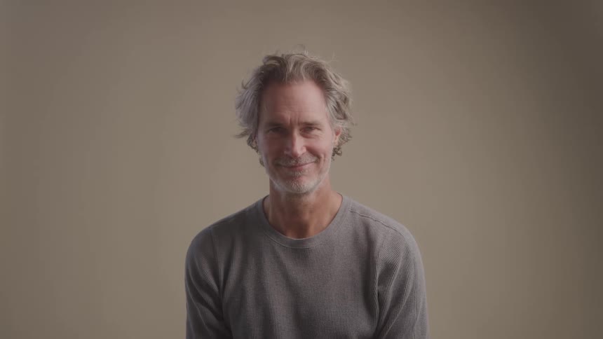 A man with gray hair and a beard smiles warmly while facing the camera in a neutral studio setting
