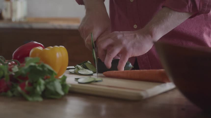 Person slices cucumber on a wooden cutting board surrounded by fresh vegetables