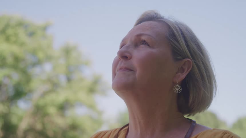 The video shows a closeup of a middleaged woman with blonde hair looking up and to the side with a serene expression she is outdoors with a clear sky and blurred green trees in the background suggesting a peaceful sunny day