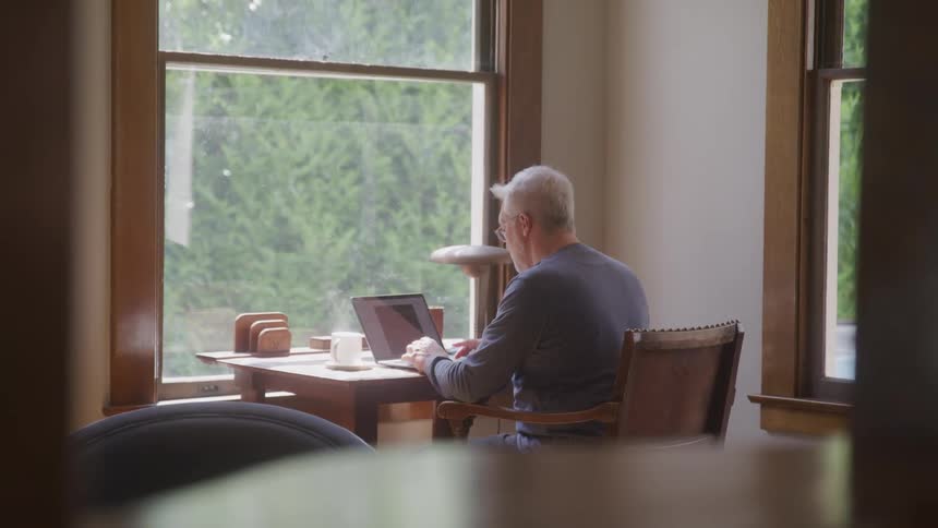 A person with gray hair and glasses is sitting in a brown armchair holding a tablet sunlight is coming through a window behind him and there are plants and a framed picture on the windowsill