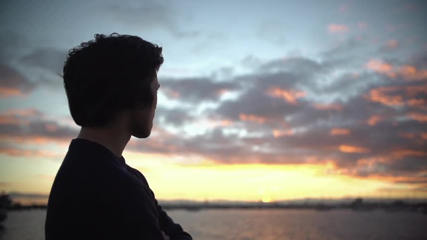 A young man gazing towards the horizon at sunset next to the ocean