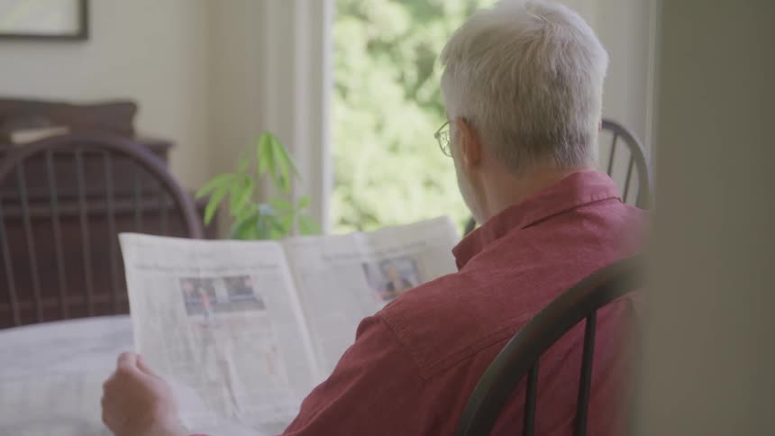A static shot from a slightly obscured angle shows an elderly person with gray hair seen from behind sitting at a table and intently reading a newspaper