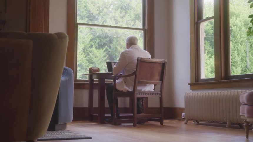 A cinematic shot from behind shows an elderly man sitting at a table with a laptop gesturing as if in a video call bathed in natural light from a large window overlooking a lush green outdoor scene