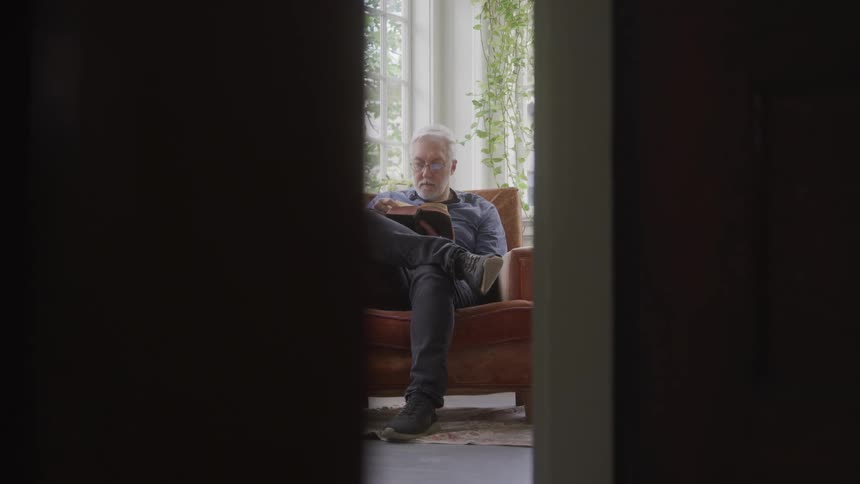 A man is partially visible reading in an armchair through a doorway in a softly lit room