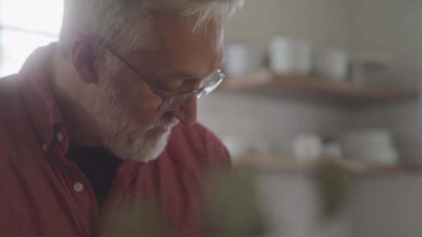A medium closeup of a man with gray hair and a beard wearing glasses and a pink shirt he is looking down and concentrating on something out of frame