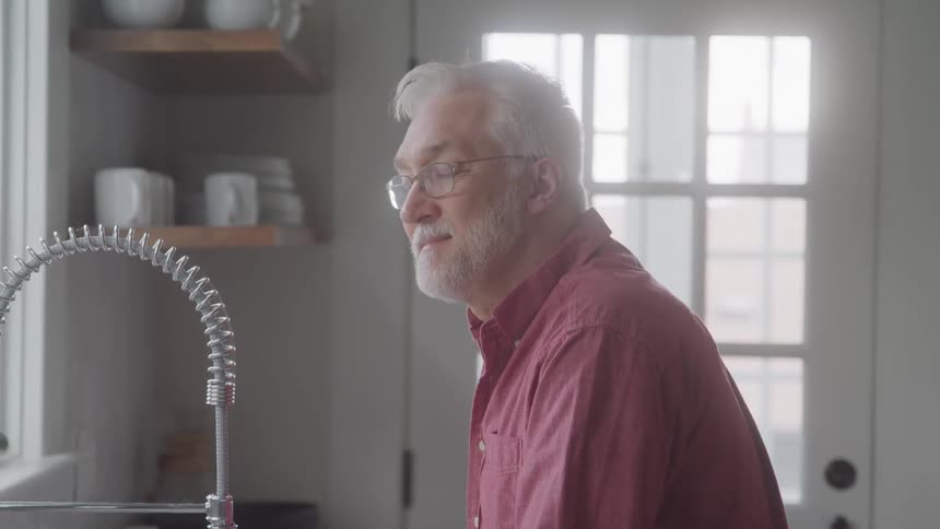 Man in red button up shirt leaning on kitchen sink looking out the window